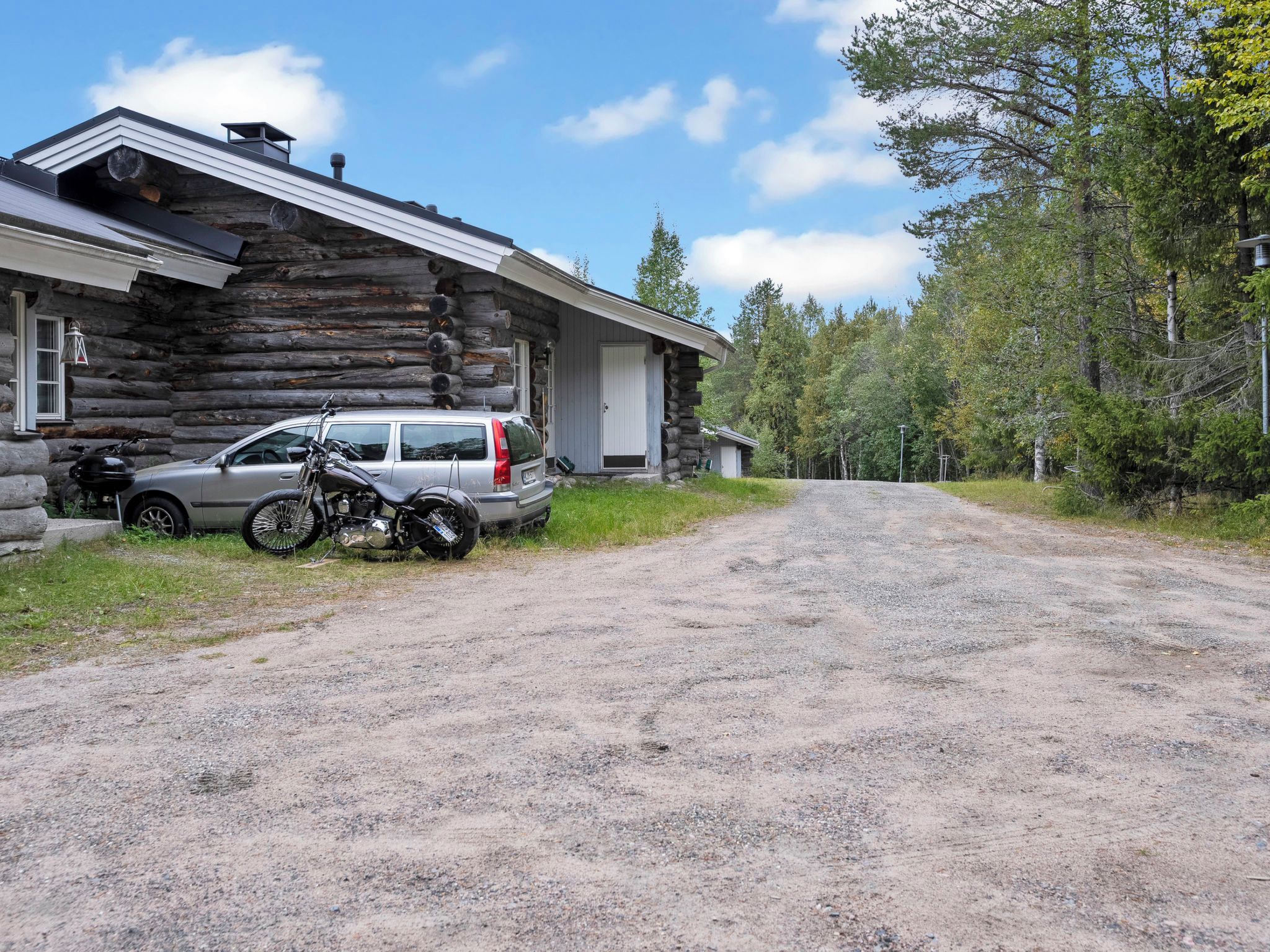 Une cabane en rondins avec une allée en gravier, une voiture garée et une moto.