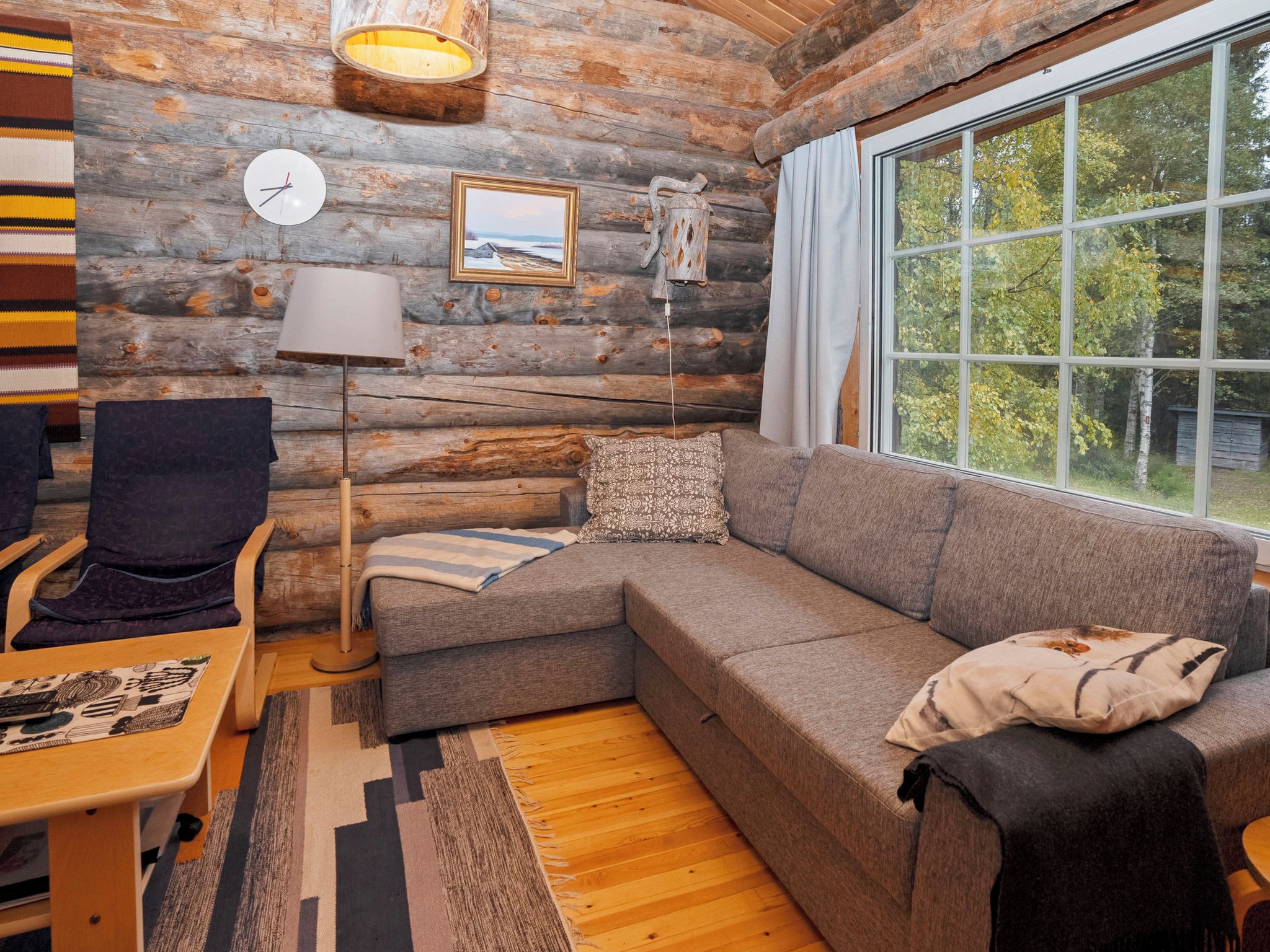 A cozy log cabin interior with a sectional sofa, a chair, a lamp, a clock, and a window with a view of trees.