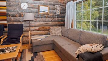 A cozy log cabin interior with a sectional sofa, a chair, a lamp, a clock, and a window with a view of trees.