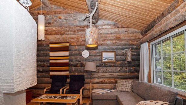Ein gemütliches Blockhaus-Interieur mit einer Couch, Sesseln, einem kleinen Tisch und einem Fenster mit Blick auf Bäume.