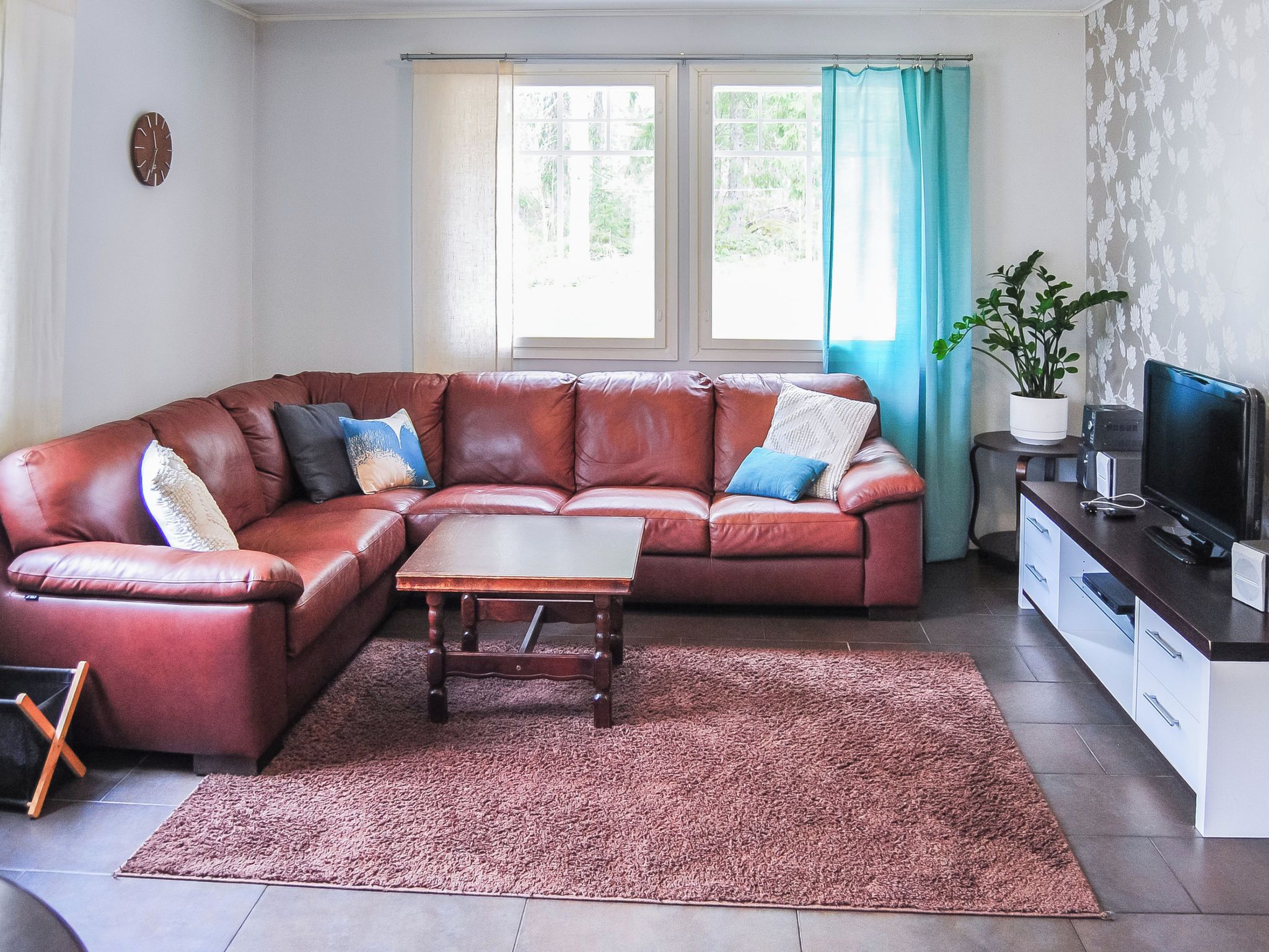 A living room with a brown leather sofa, a wooden coffee table, a large window with curtains, a potted plant, and a television on a stand.