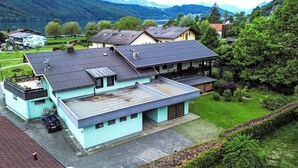 Un bâtiment moderne à deux étages, doté d’un toit plat et d’un balcon, entouré d’une végétation luxuriante et surplombant un paysage montagneux.