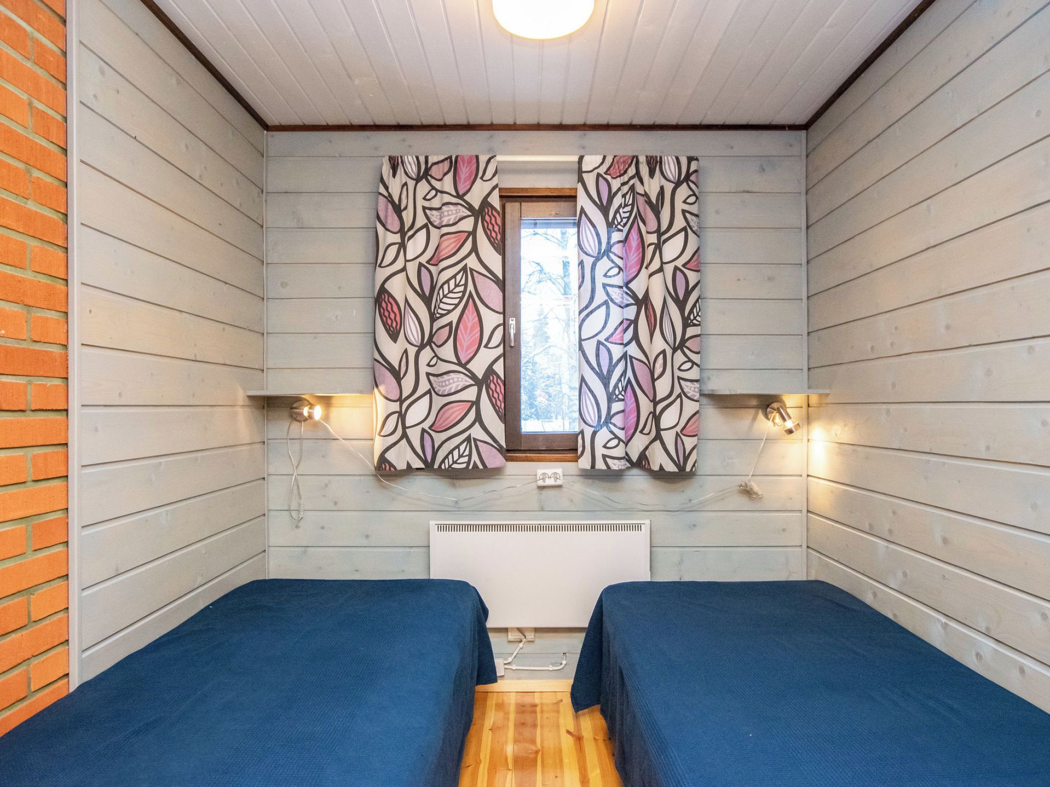 A room with two beds, a window with patterned curtains, a radiator, and wall-mounted lights.