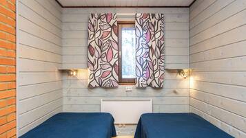 A room with two beds, a window with patterned curtains, a radiator, and wall-mounted lights.