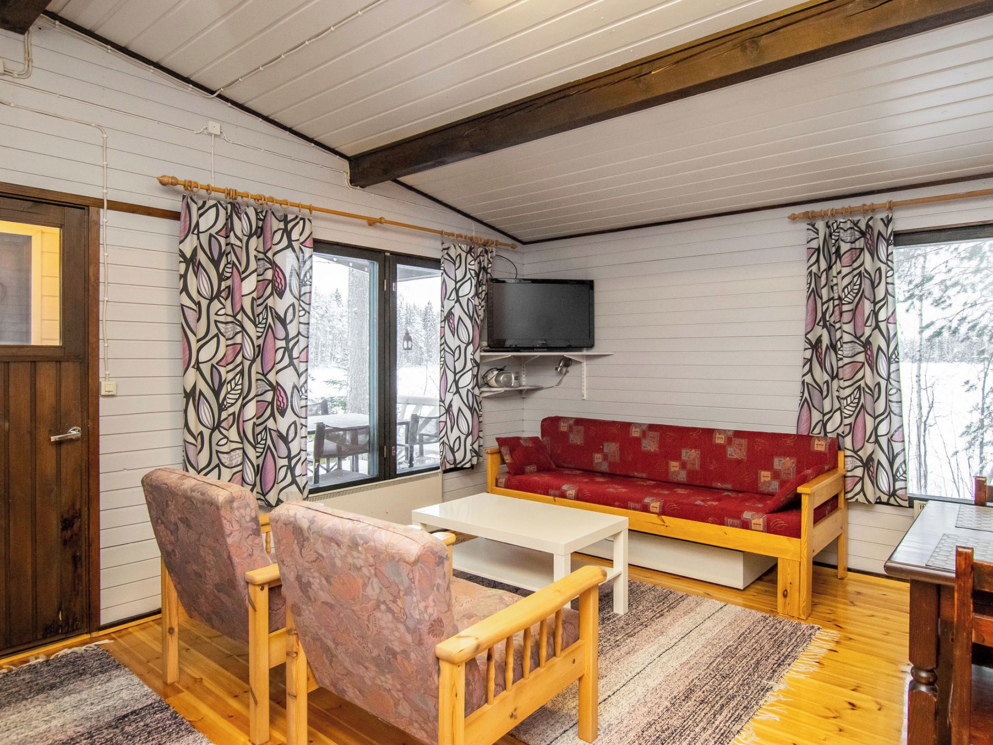 Una sala de estar acogedora con un sofá rojo, sillas de madera, un televisor y una ventana con cortinas estampadas.