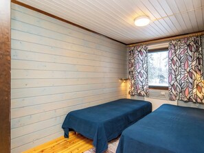 A room with two beds, wooden walls, a window with patterned curtains, and a ceiling light.