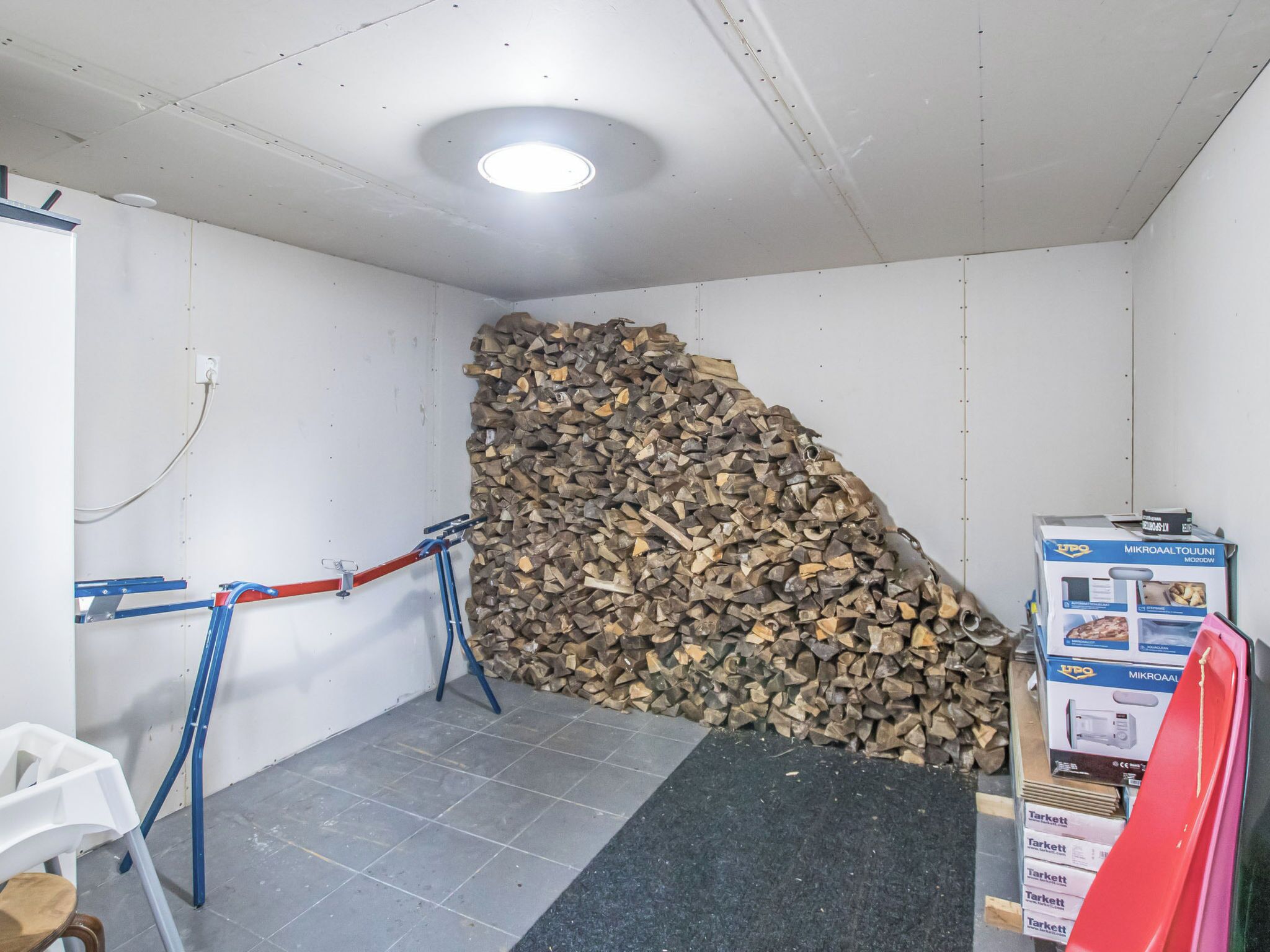 Ein Zimmer mit einem Holzstapel an der Wand, einem weißen Stuhl, einer blau-roten Leiter und einer mit ‚Tarkett‘ beschrifteten Kiste auf dem Boden.