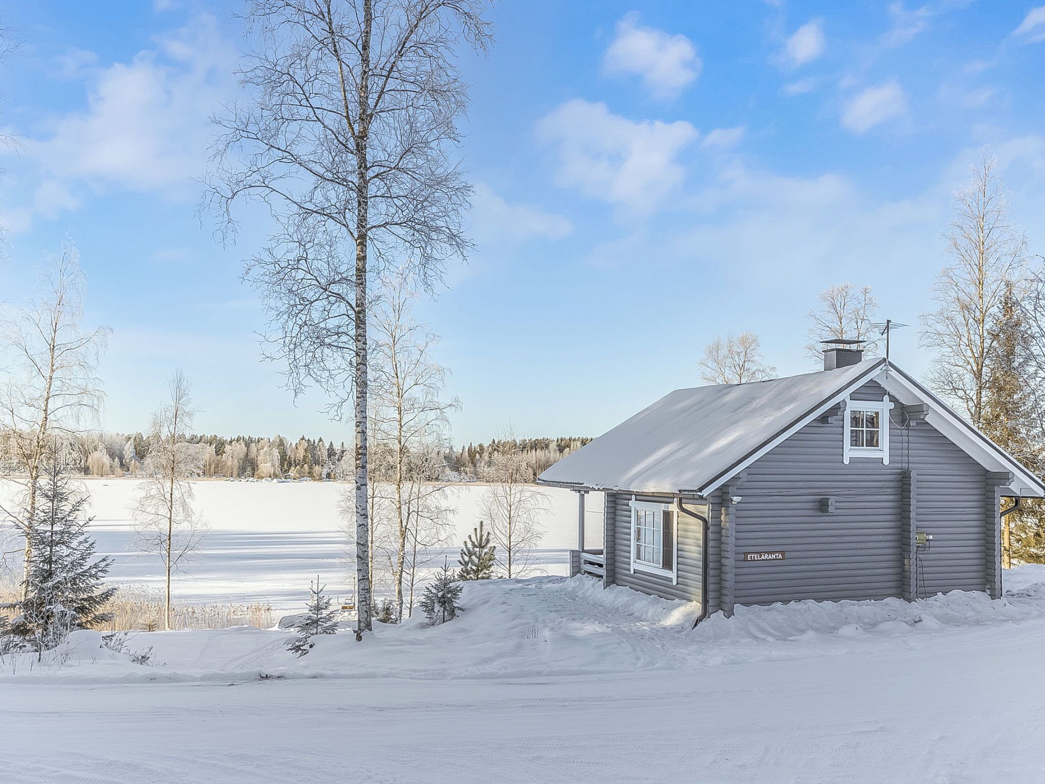 A small, single-story cabin with a gabled roof and a chimney, surrounded by snow-covered ground and sparse trees.