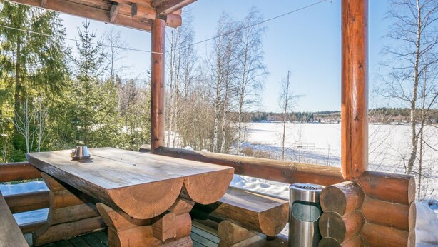 A wooden deck with a table and benches, a metal trash can, and a view of a snowy landscape with trees. - Eteläranta by Interhome (Joutsa)