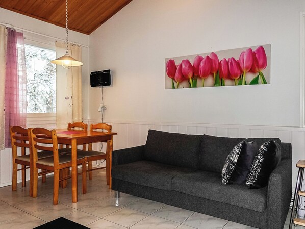 Una sala de estar con una mesa de comedor, sillas, un sofá, un televisor montado en la pared y un ventanal con cortinas.