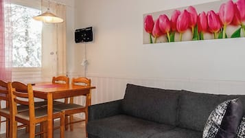 Tunnelmallinen olohuone, jossa on tummanharmaa sohva, puinen ruokapöytä tuoleineen, ikkuna verhoilla ja seinälle asennettu televisio.