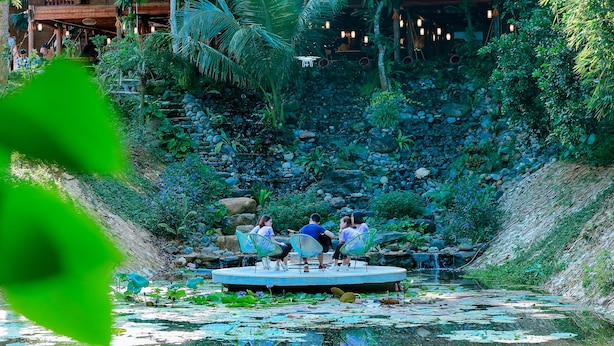 A group of people seated on chairs around a pond with a rocky backdrop and lush greenery. - May Tropicall Villas (Hanoi)