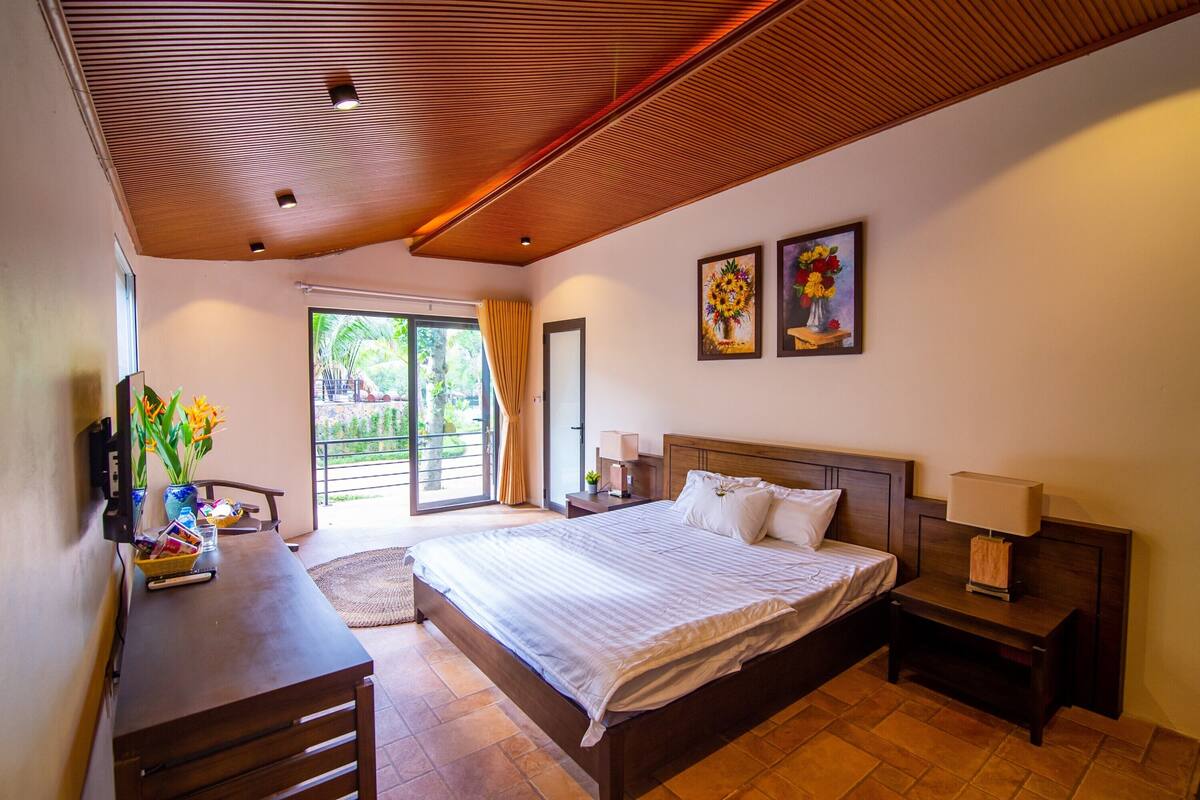 A hotel room with a large bed, a desk, a chair, a TV, and a window with a view of greenery.