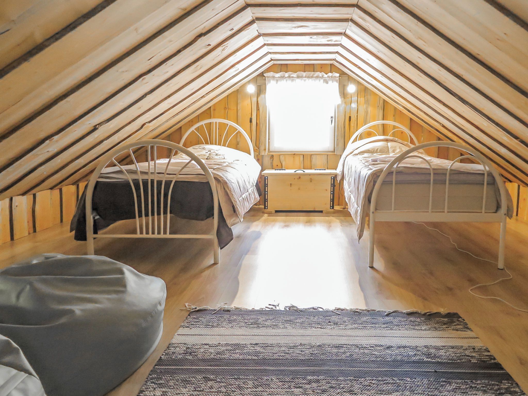 Quarto de sótão pequeno com duas beliches de metal, uma janela e piso de madeira.