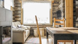 A cozy log cabin interior with a fireplace, a sofa, a dining table, and a large window offering a view of the snowy outdoors.