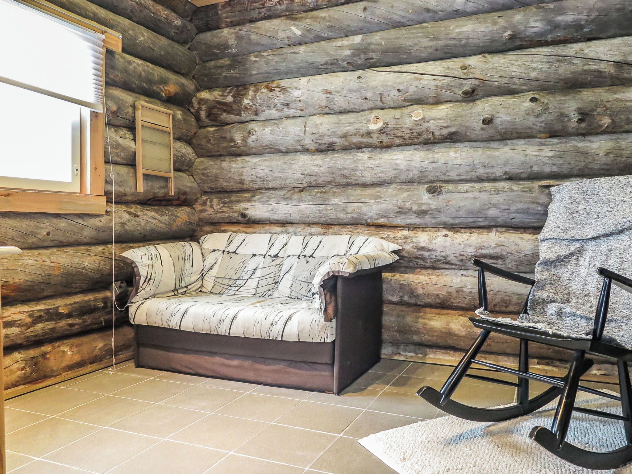 Interior de una cabaña de troncos con un sofá, un sillón mecedor y una ventana con persianas.