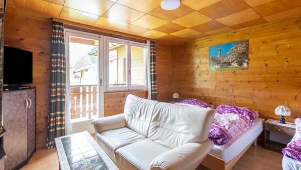 Una habitación acogedora con techo de madera, un sofá, una cama con manta púrpura, una mesita y un televisor sobre un mueble.