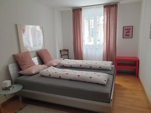 Een slaapkamer met een bed, kussens, een stoel, een nachtkastje, een rode bank en een muur met een bordje.