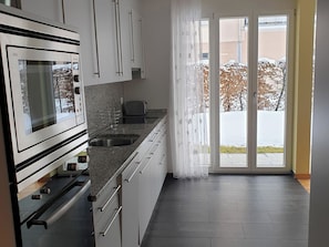 Een moderne keuken met een ingebouwde oven, een spoelbak en een broodrooster. Er is een schuifpui die leidt naar een balkon met uitzicht op bomen en een gebouw.