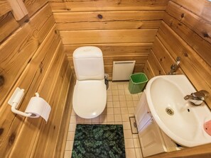 Une salle de bain rustique, aménagée dans une cabane en bois, avec des toilettes, un lavabo et une poubelle verte.