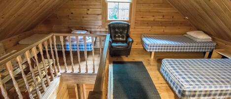 A loft bedroom with a wooden ceiling, a black leather chair, two mattresses, a small table, and a window.