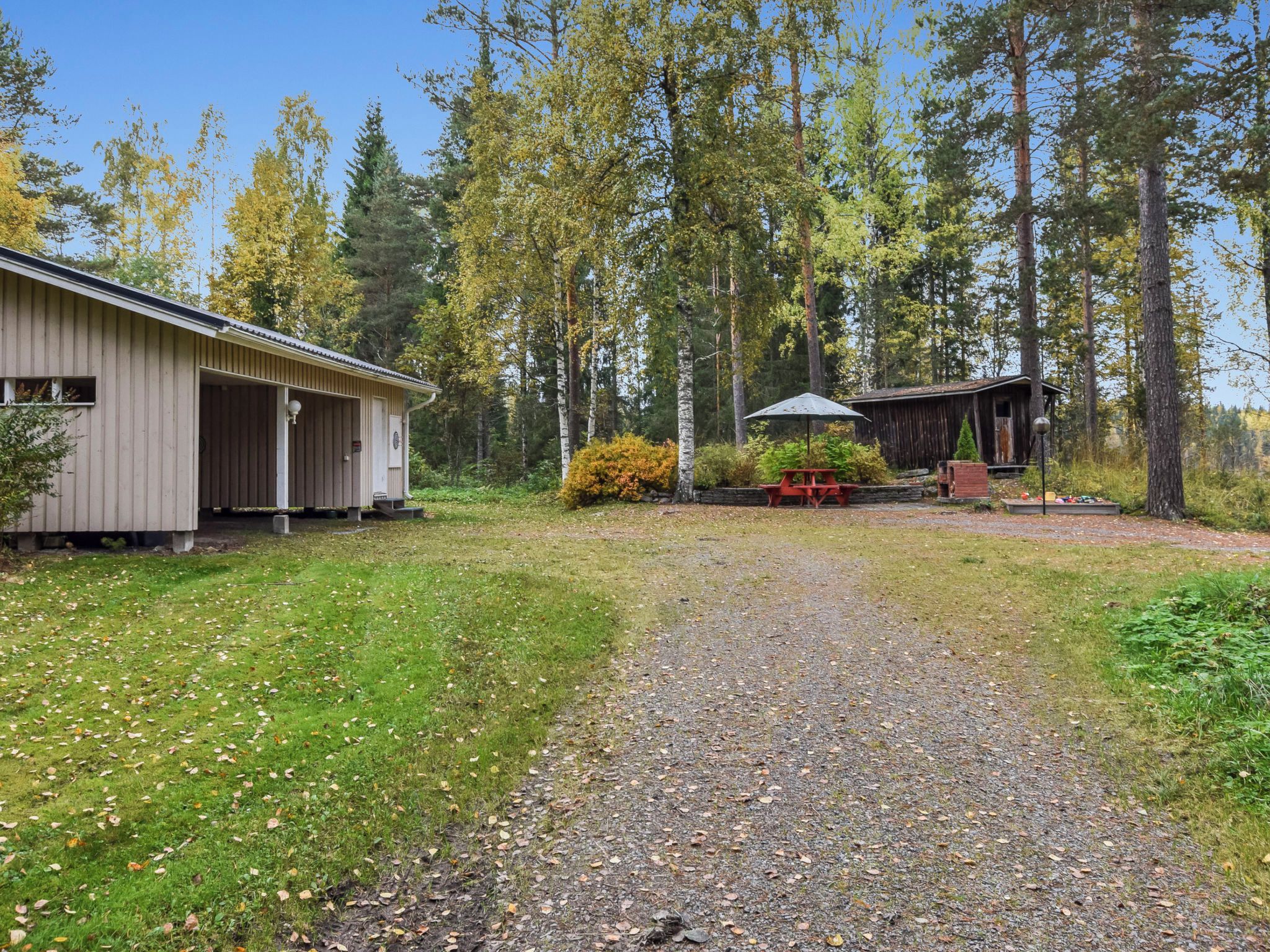 Un chemin de gravier mène à un chalet en bois, avec un abri de jardin et une table de pique-nique, le tout situé dans une zone boisée.