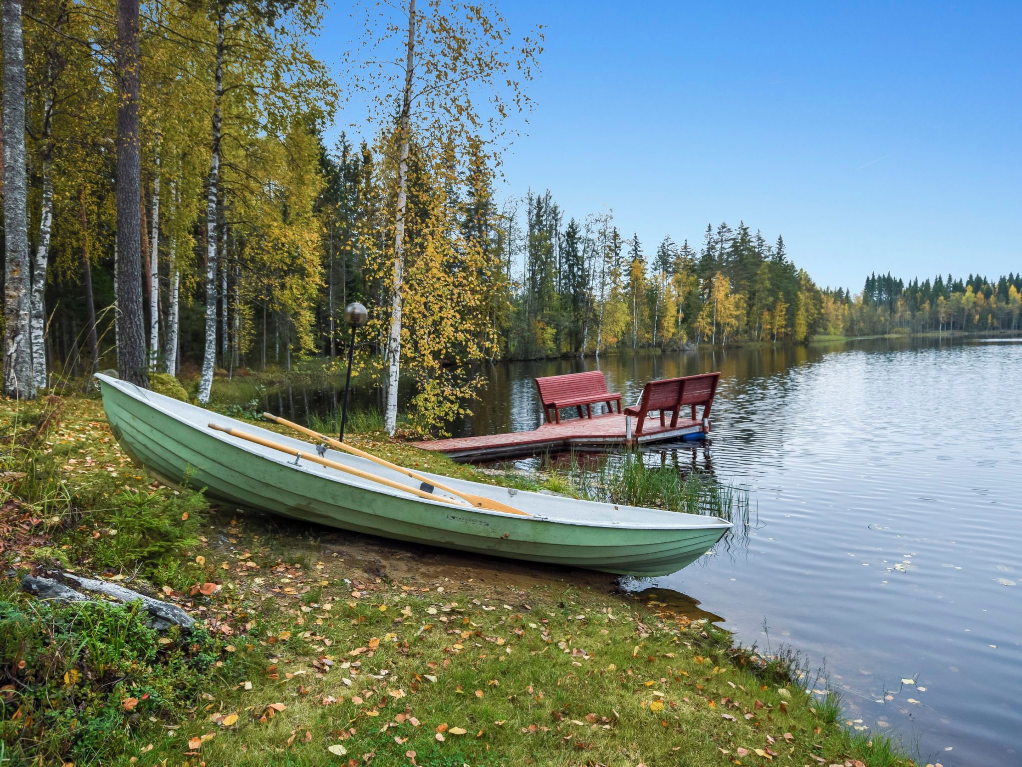 A calm lake with a green rowboat and a dock with red chairs.