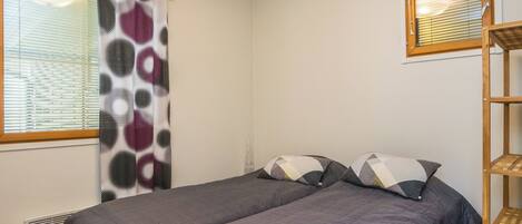 Une chambre avec deux lits, une fenêtre avec des stores, un radiateur et une étagère en bois.