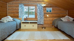 Ein gemütliches Cabin-Zimmer mit zwei Betten, Holzbalkendecke, Fenster mit Vorhängen, Schreibtisch mit Lampe und einem gerahmten Bild an der Wand.