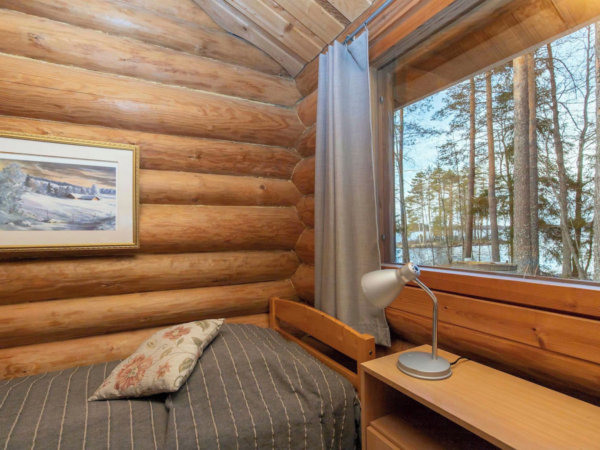 Habitación tipo cabaña de madera con una cama, un escritorio, una lámpara, un ventanal con vista a árboles y un cuadro en la pared.