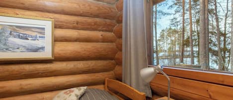 A log cabin room with a bed, a desk, a lamp, a window with a view of trees, and a framed picture on the wall.