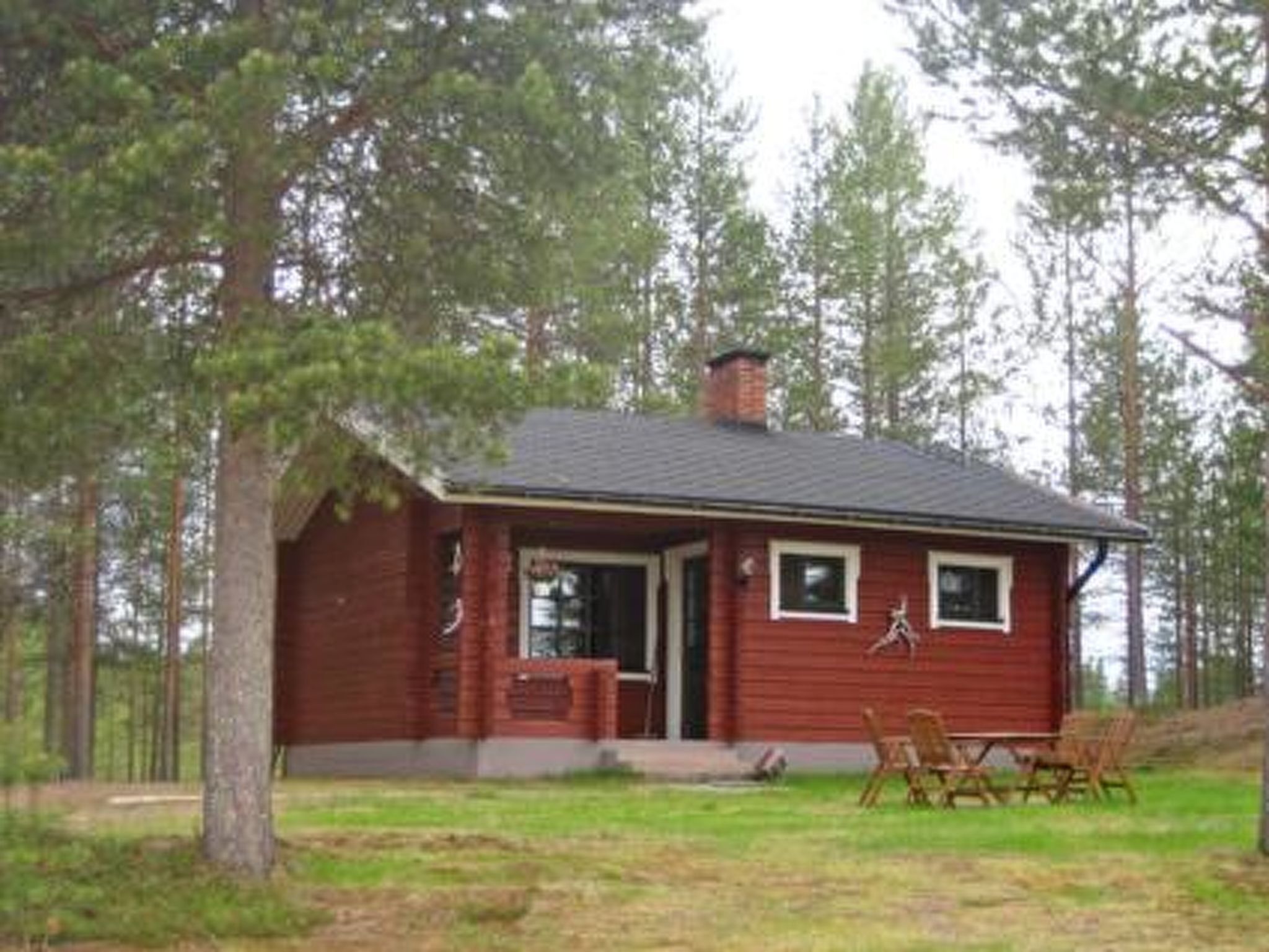Une petite cabane à un seul étage, avec une façade rouge, une cheminée et une véranda à l’avant, comprenant deux chaises et une table.