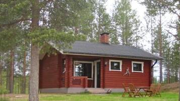 A small, single-story cabin with a red exterior, a chimney, and a front porch with two chairs and a table.