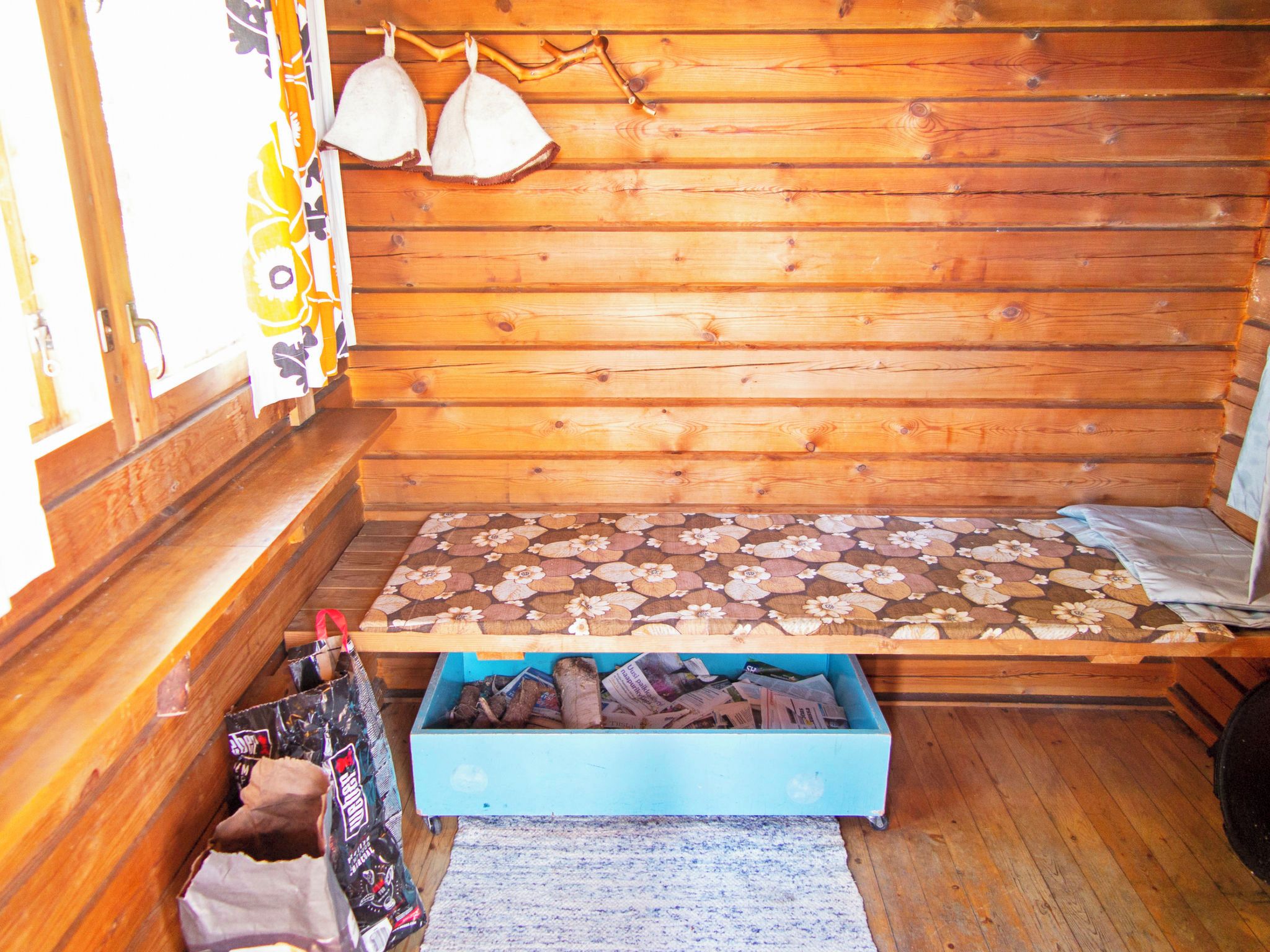 A wooden cabin interior with a bed, a blue storage box, and a window with curtains.
