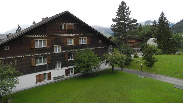 A two-story wooden building with multiple windows and a sloped roof, surrounded by a grassy area and trees.