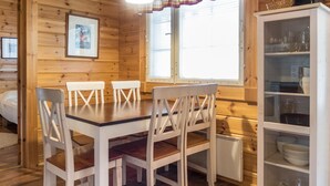 Ein Holzblockhaus-Interieur mit Esstisch, vier Stühlen, einem Schrank mit Geschirr und einem Fenster mit Jalousien.