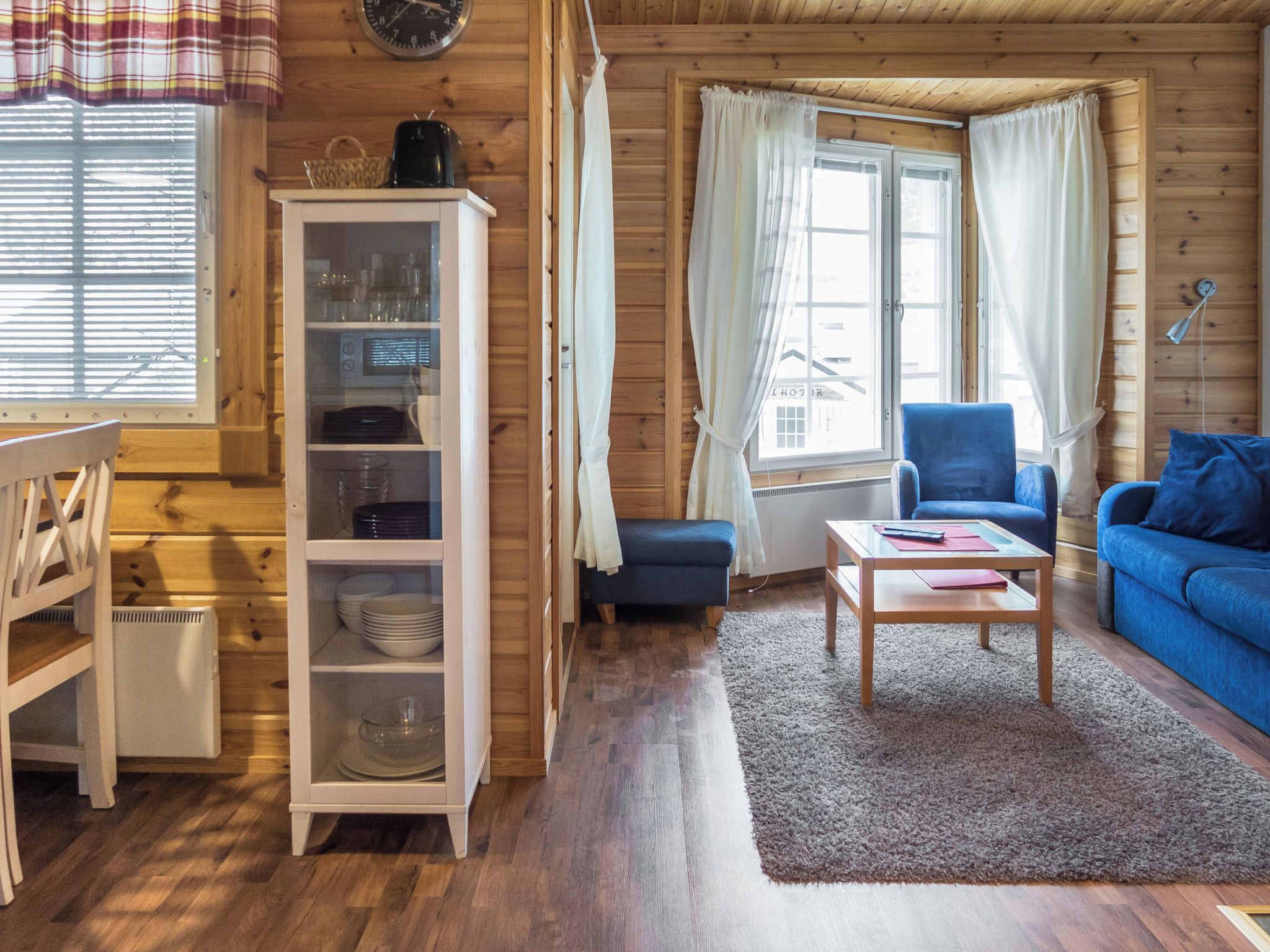 Een gezellig interieur van een blokhut met een eettafel, een blauwe bank, een kast met servies en een raam met gordijnen.