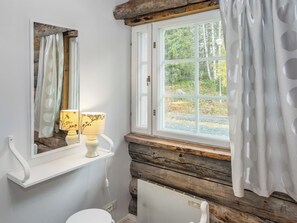 Un baño con pared de tronco de madera, un radiador blanco, una ventana con vistas a árboles y dos lámparas en un estante.