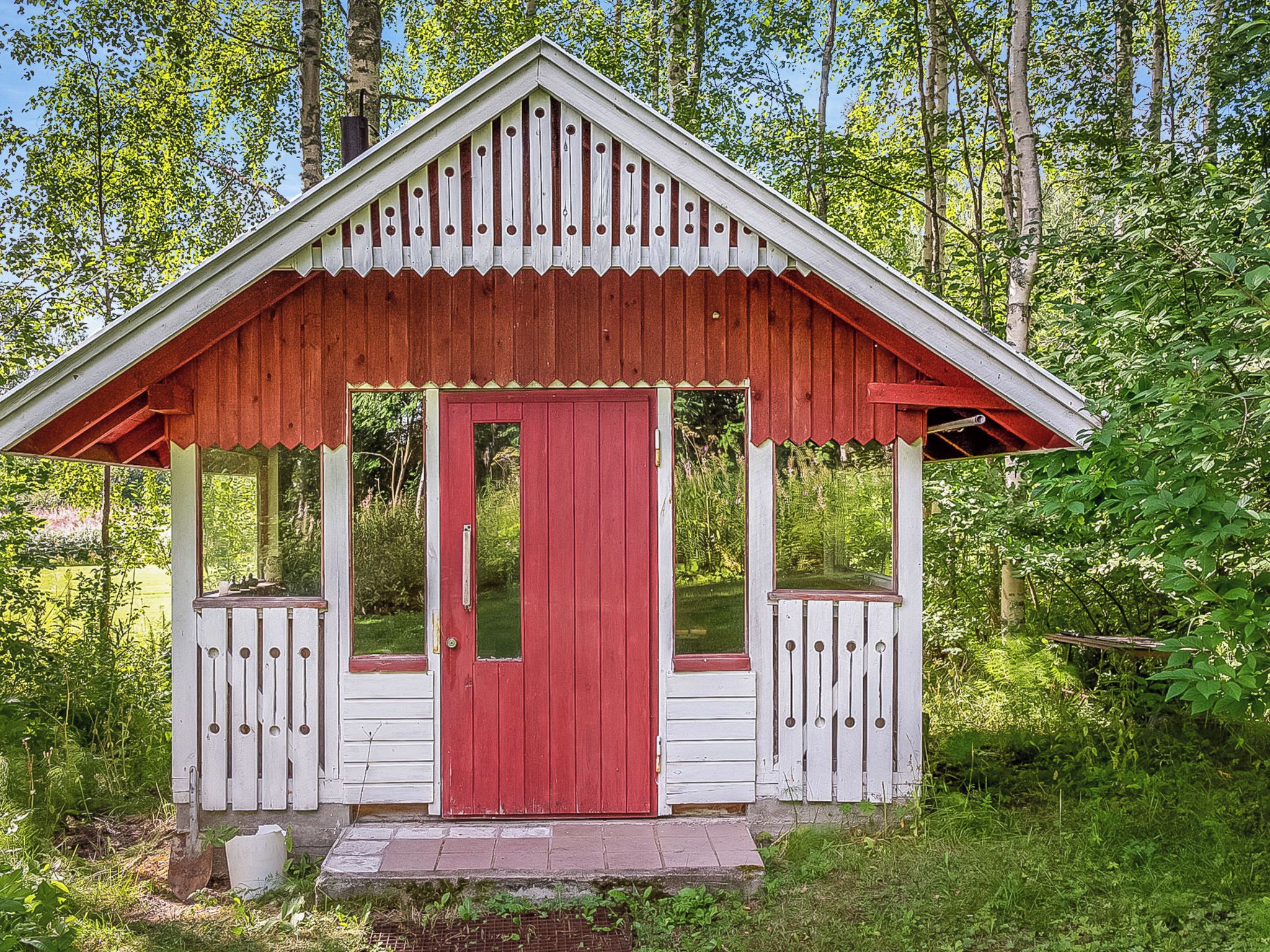Une petite construction en bois d’un seul étage, avec une porte rouge et des boiseries blanches, entourée d’une végétation luxuriante.