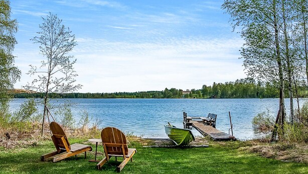 Un cadre idyllique au bord d’un lac, avec deux chaises en bois et une table sur une pelouse, une jetée avec un bateau, et des arbres qui bordent le rivage.
