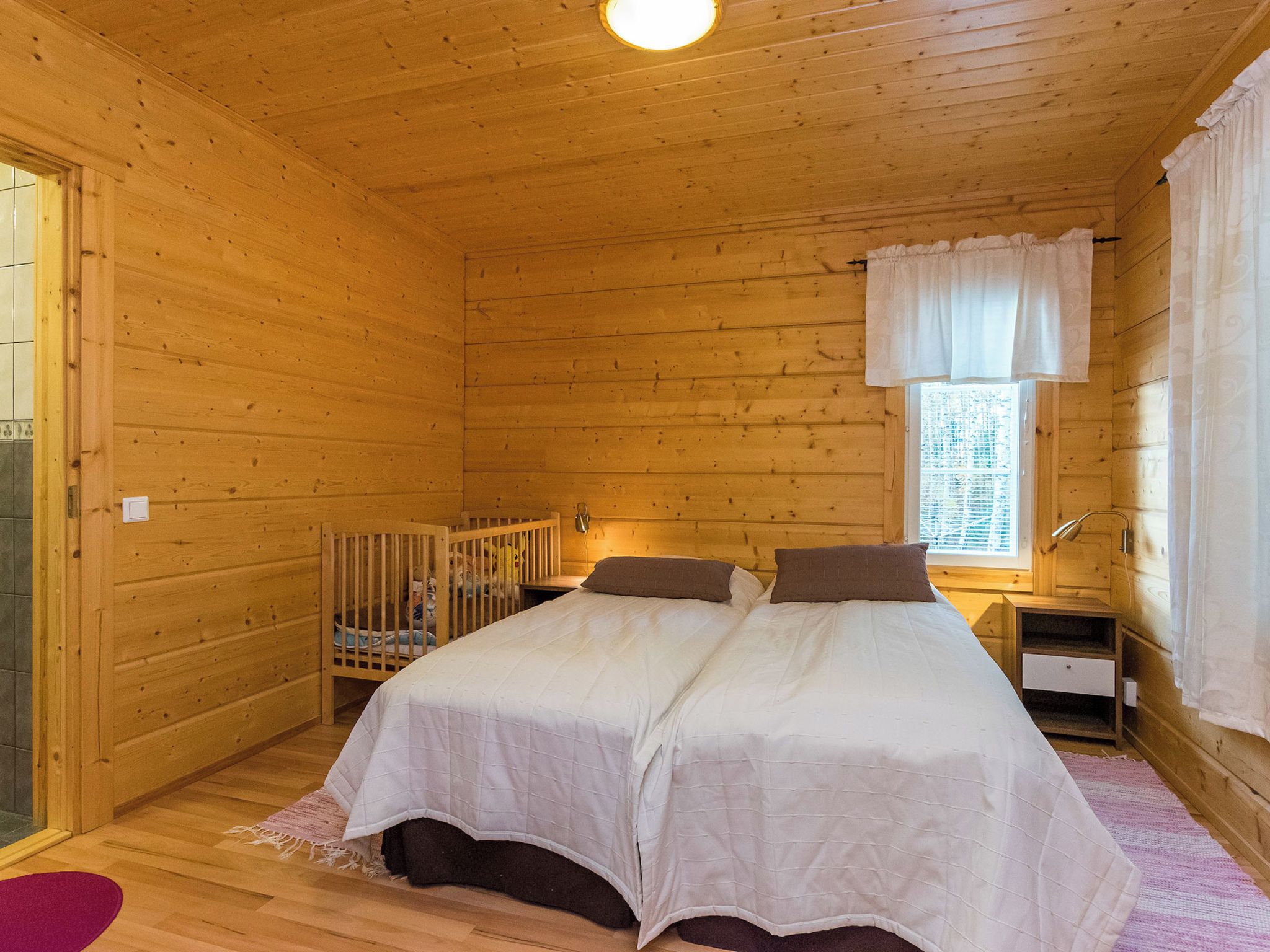 Een slaapkamer in een houten hutje, met een bed, ledikant, raam met gordijnen en nachtkastje.
