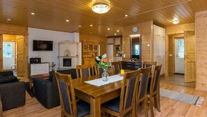 Un interior de madera con una mesa de comedor dispuesta para seis comensales, una chimenea, un televisor montado en la pared y una zona de cocina visible a través de una puerta abierta.
