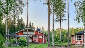 Une cabane rouge dotée d’un porche et d’un espace extérieur aménagé, entourée de grands arbres et d’une pelouse bien entretenue.
