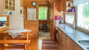 Une pièce confortable, aménagée comme un chalet, avec des murs et des planchers en bois, une table à manger en bois, une cuisine équipée d’un évier et de placards, et une fenêtre donnant sur un paysage verdoyant.