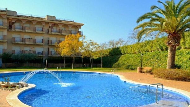 Una piscina con fuente, rodeada por un patio de ladrillo, con un edificio y palmeras al fondo.