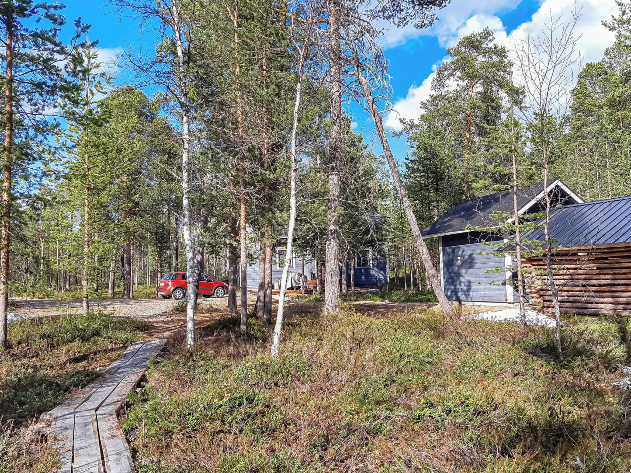 Une zone boisée avec un chalet en bois, une voiture rouge et un chemin pavé.