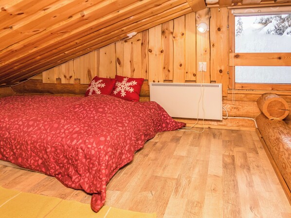 A bedroom with a sloped ceiling, wooden walls, a bed with red bedding, and a window with a view of snow-covered trees.