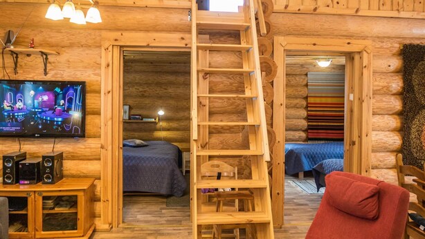 A log cabin interior with a wooden ladder leading to a loft, a television, and a bed with a blue blanket.