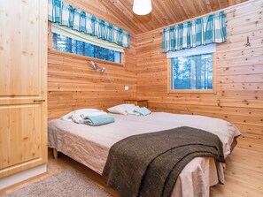 Ein Schlafzimmer in einer Holzhütte mit einem Bett, zwei mit Vorhängen drapierten Fenstern, einem Nachttisch und einem Holzschrank.