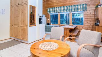 A cozy cabin interior with a dining area, wooden cabinets, and a view of the forest through the windows.